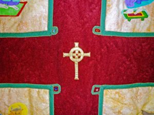 Gold fabric Celtic cross with embedded gemstone on a burgundy back ground in the center of a quilt depicting the Lindisfarne Gospels