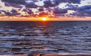 Solar eclipse occurring at sunset over dark ocean waves and a sandy shore with clouds filling the dusk sky above the horizon.