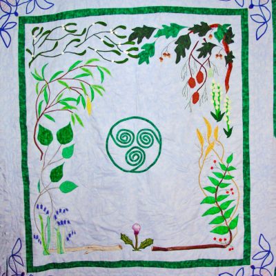 Quilt depicting a central triskele design, surrounded by various plants and flowers